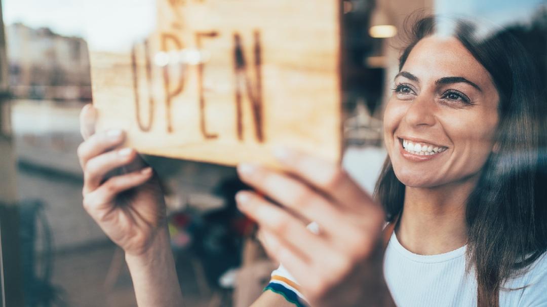 A smiling woman holding an "open" sign in a shop window, reflecting the street outside.
