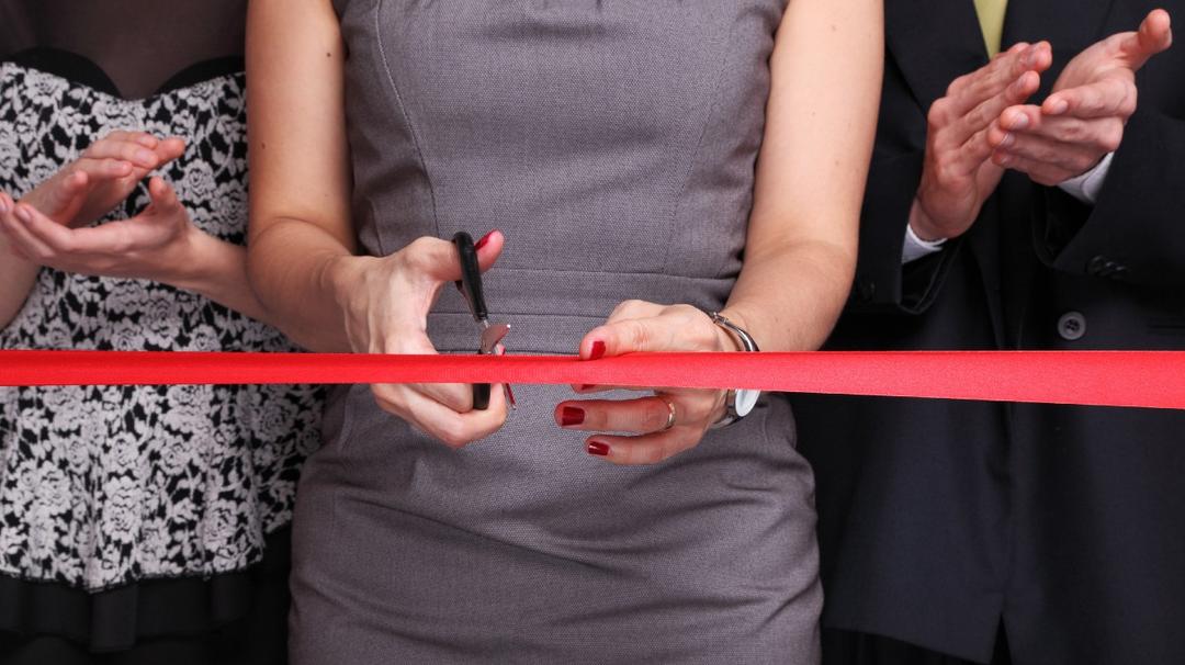 A woman in a gray dress cuts a red ribbon with scissors, standing between two applauding people partially visible in the frame.